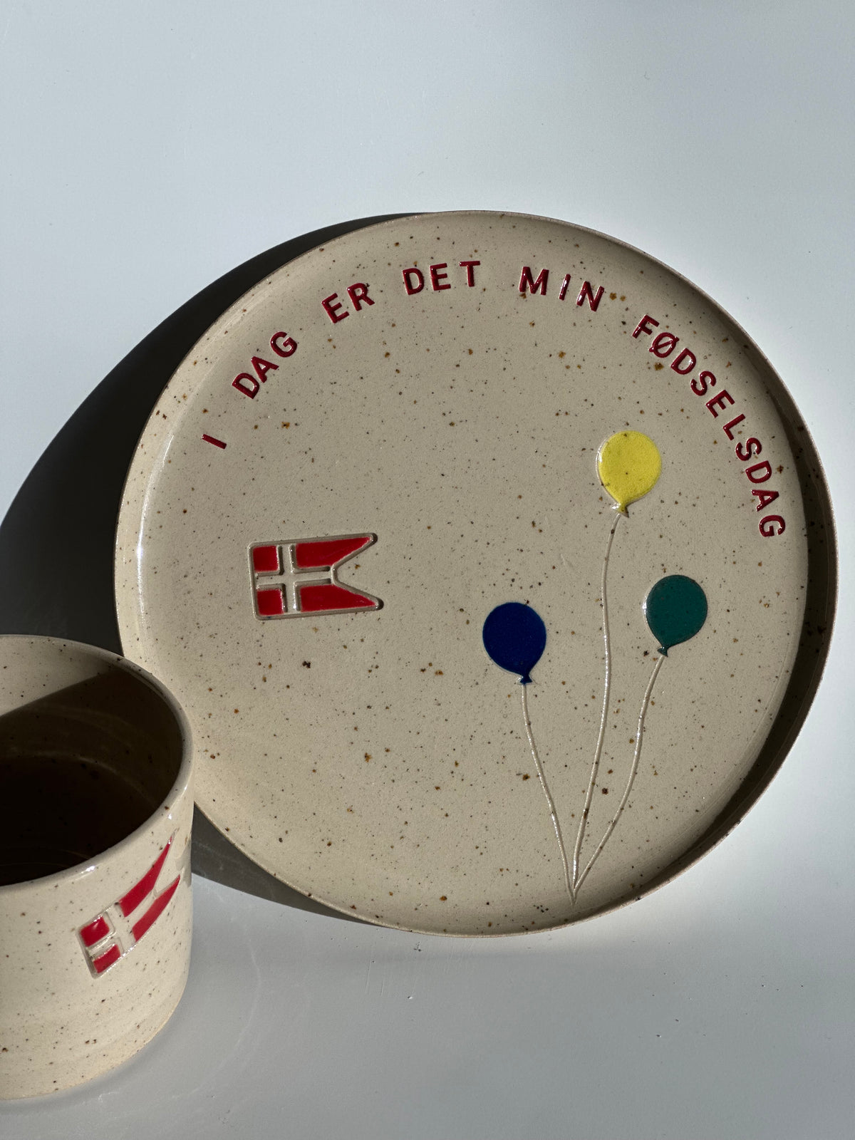 Fødselsdagstallerken i keramik med teksten I dag er det min fødselsdag, Dannebrog og tre balloner, med koppen ved siden af